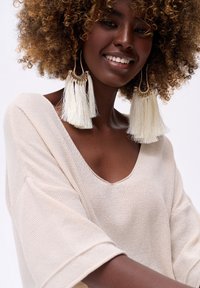 Lachende vrouw met krullend haar die grote witte franjes oorbellen draagt en een beige gebreide trui met een brede V-hals.