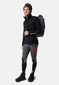 Schwarze Jacke mit "Rossignol"-Logo, graue Leggings mit rotem Emblem und schwarze Stiefel. Schwarz-weißer Rucksack mit Muster.