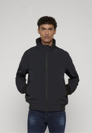 Mężczyzna ubrany w czarną kurtkę na zamek z wysokim kołnierzem i rękami w kieszeniach, w ciemnoniebieskich dżinsach, stoi na jednolitym jasnym tle.