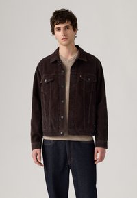 Donkerbruine corduroy jas met een klassieke kraag, knoopsluiting aan de voorkant, twee borstzakken en een rechte snit. Gecombineerd met een beige trui en donkere spijkerbroek.