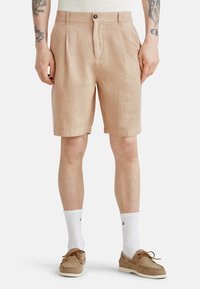 Pantaloni corti beige a fantasia realizzati in un tessuto strutturato, con pieghe, chiusura con bottone frontale e tasche laterali. Indossati con calzini bianchi e mocassini.