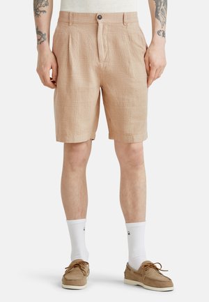 Pantalones cortos de color beige con un patrón, hechos de una tela texturizada, con plisados, cierre de botón frontal y bolsillos laterales. Llevados con calcetas blancas y mocasines.