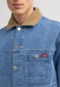 Denimtakki, jossa on beige corduroy-kaulus, hopeiset napit ja etutasku. Vasemmassa taskussa on punainen tuotemerkki.