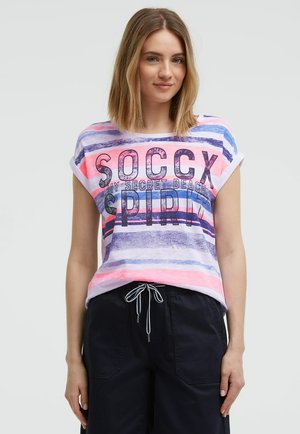 Junge Frau mit schulterlangem blondem Haar, die ein gestreiftes Shirt mit Text und dunkle Kordelhose trägt, steht vor einem hellen Hintergrund.