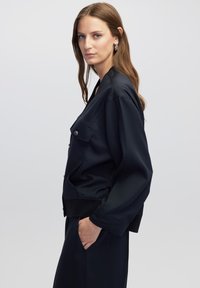 Chaqueta azul marino fabricada en tejido suave, con un bolsillo frontal, corte relajado y hombros caídos. Combinada con un fondo a juego.