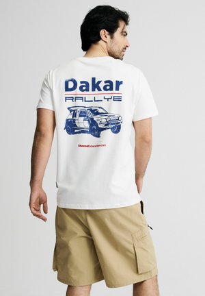 Mężczyzna w białej koszulce z niebieskim napisem „Dakar Rallye” i grafiką rajdowego samochodu na plecach, w połączeniu z beżowymi szortami typu cargo.