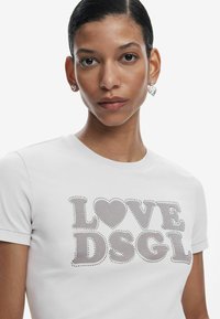 Maglietta bianca a maniche corte con testo decorato da borchie argento che recita "LOVE" e "DSGL", con una forma a cuore nel design. Tessuto liscio.