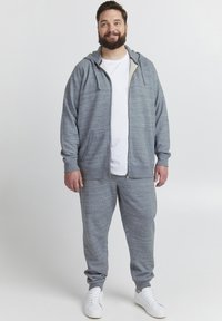 Lys grå hettejakke med glidelås og kontrastfôr, sammen med matchende joggebukser. Har en myk tekstur og sidelommer. Hvite joggesko.