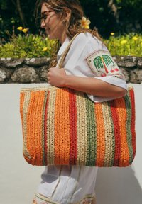 Sac fourre-tout tissé avec des motifs rayés orange, vert et rouge. Fabriqué en fibres naturelles avec des poignées robustes. Couture détaillée le long des bords.