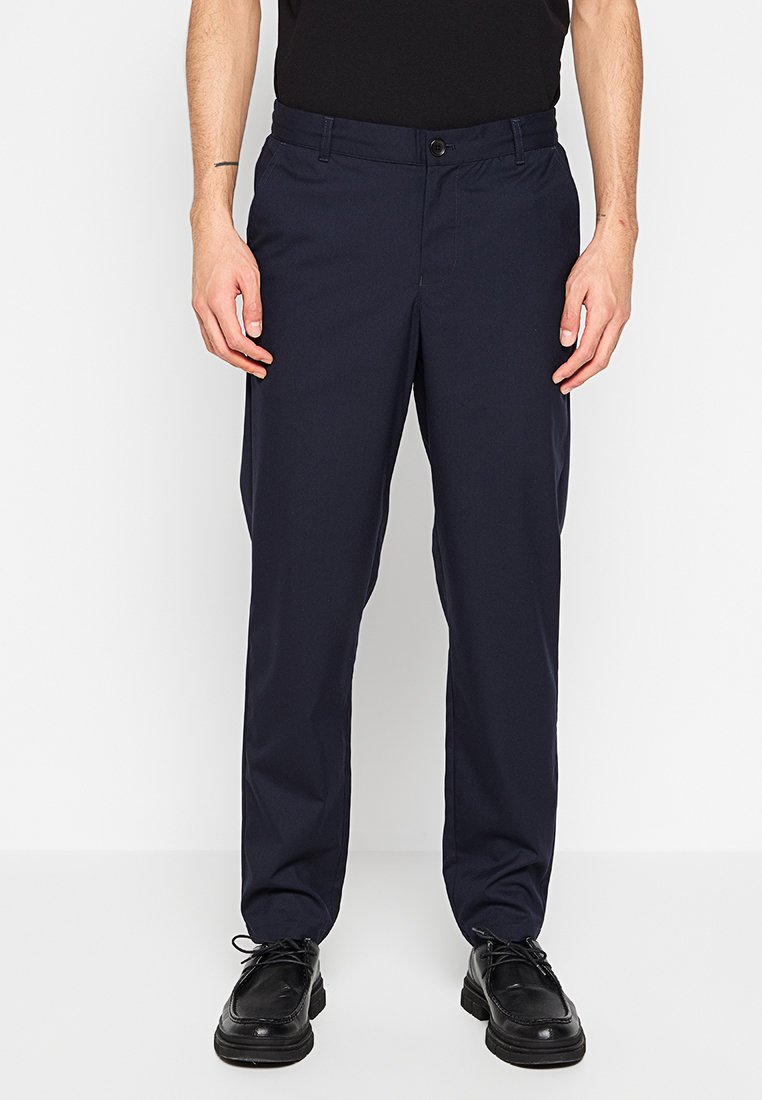 Marineblaue Hose mit geradem Schnitt, aus leichtem Stoff gefertigt, mit Knopfverschluss und Gürtelschlaufen, kombiniert mit schwarzen Schuhen.