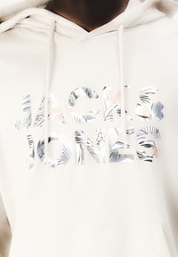 Haut à capuche crème avec le texte "JACK & JONES" en imprimé floral texturé et coloré. Comprend une capuche avec cordon de serrage et une poche frontale. Tissu doux.