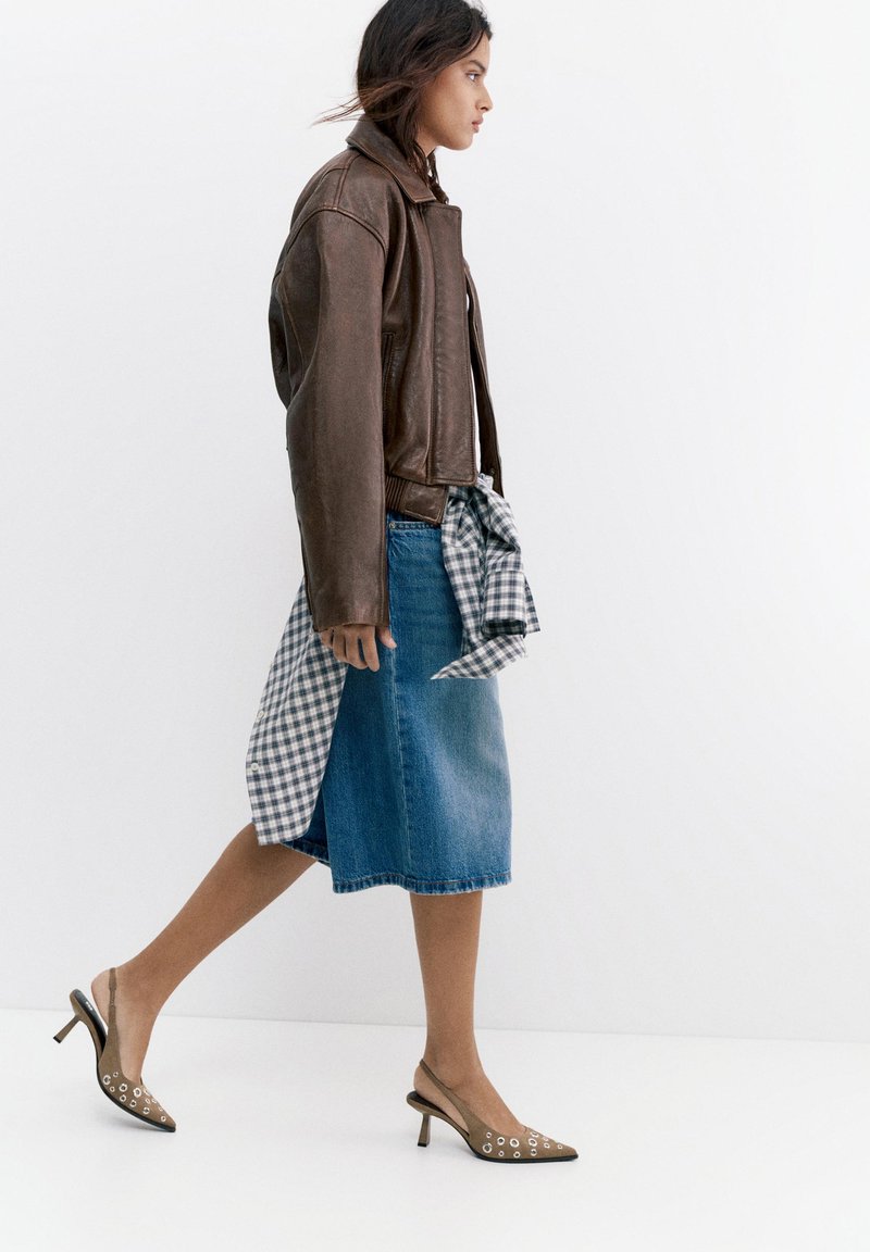 Femme marchant portant une veste en cuir marron, une jupe en jean avec une chemise à carreaux nouée, et des talons slingback marron avec des détails œillet.