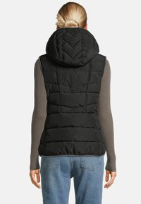 Frau mit blonden Haaren im Dutt, die eine schwarze gesteppte Kapuzenweste über einem grauen, gerippten Langarmoberteil und blauen Jeans trägt und von hinten blickt.