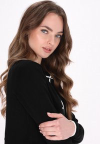 Maglione nero con dettagli e bottoni bianchi. La modella ha capelli ondulati e castani ed è posizionata in modo da mostrare i dettagli del polsino e del colletto del maglione.