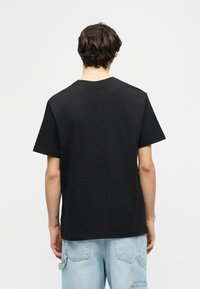 Άτομο με κοντά σκούρα μαλλιά που φοράει μια μονόχρωμη μαύρη κοντομάνικη μπλούζα και ανοιχτόχρωμο τζιν facing away σε λευκό φόντο.