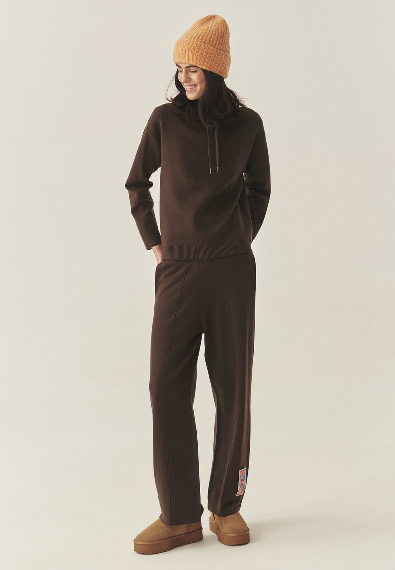 Pruun kudumissviiter kapuutsiga, ühendatud sobivate kitsenevate pükstega. Mõlemal on pehme tekstuur. Pruunad sussid ja tekstureeritud oranž beanie.