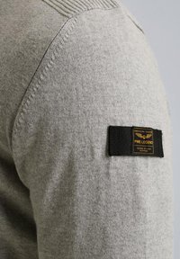 Grauer Strickpullover mit geripptem Kragen und strukturiertem Stoff; verfügt über ein rechteckiges schwarzes Etikett mit goldener Schrift auf der linken Schulter.