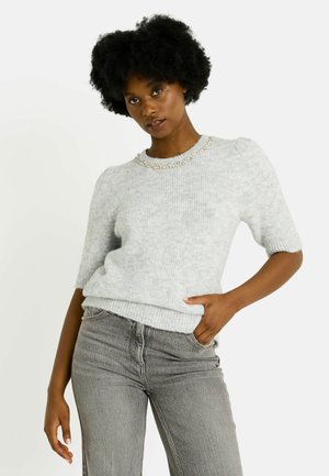 Suéter de punto gris claro con mangas cortas abullonadas y dobladillo acanalado, con un detalle de cuentas en el cuello. Combinado con jeans grises.
