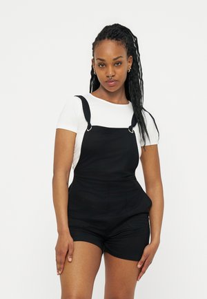 Jonge vrouw met een wit shirt met korte mouwen en zwarte korte tuinbroek, staand tegen een effen lichte achtergrond.