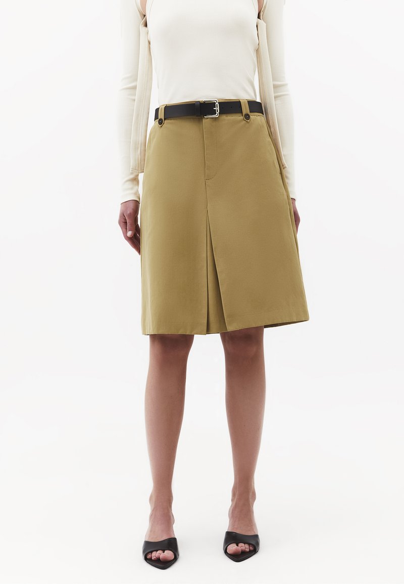 Mujer con falda beige hasta la rodilla con pliegue frontal y cinturón negro, combinada con una camiseta blanca de manga larga y tacones abiertos negros.