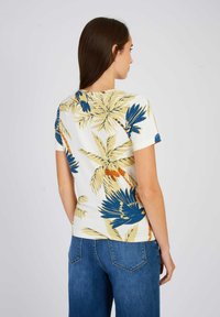 Chemise à manches courtes blanche avec un imprimé de feuilles tropicales en jaune et bleu. Tissu doux, coupe ajustée et col rond.