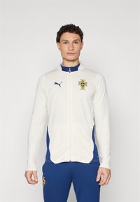 Giacca da uomo con zip color crema e dettagli blu. Presenta il logo Puma e il crest del Portogallo. Maniche lunghe, texture liscia, vestibilità atletica.