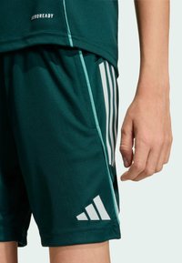 Pantalones cortos deportivos verdes con logo blanco de Adidas y diseño de tres rayas. Hechos de tela ligera y suave, con un ajuste relajado y bolsillo lateral.