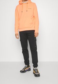 Felpa arancione neon con tasca a marsupio frontale e cordini neri, abbinata a pantaloni cargo neri con tasche con zip e dettagli.