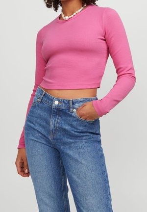 Personne portant un haut court rose à manches longues, un jean bleu taille haute et un grand collier de perles, main dans la poche du jean.
