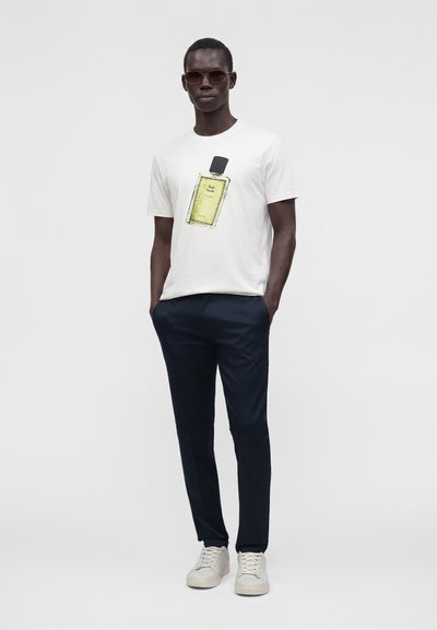 Homme portant un t-shirt blanc avec un motif de bouteille de parfum, pantalon bleu foncé, baskets blanches et lunettes de soleil, debout les mains dans les poches.