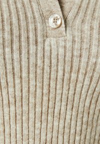 Lichtbeige geribde trui met een V-hals en een enkele ronde, doorzichtige knop. De textuur lijkt zacht en gebreid met contrasterende strepen.