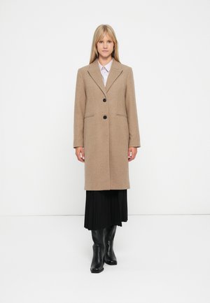 Beige ullblandede frakk med en strukturert design, notched krage, to svarte knapper og sidelommer, parret med et svart plissert skjørt og knelengde støvler.