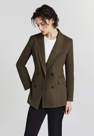 Donna che indossa un blazer doppiopetto di un verde oliva scuro sopra una camicia bianca e pantaloni neri, con capelli corti e ricci, che guarda verso il basso a sinistra.
