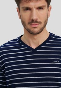 Camiseta de manga corta con cuello en V a rayas en azul marino y gris, con un pequeño logo en el lado izquierdo. La tela parece suave con una ligera textura.