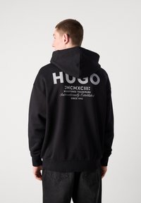 Černá mikina s kapucí a velkým bílým logem "HUGO" na zadní straně, které obsahuje další text. Design zahrnuje kapuci a vroubkované cuffs.