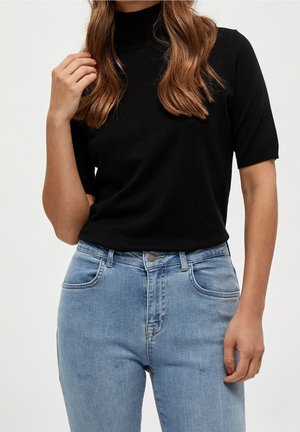 Femme aux cheveux bruns ondulés portant un pull à col roulé noir à manches courtes et un jean taille haute bleu clair, posant avec une main près des cheveux.