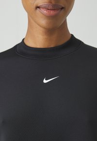 Partie inférieure du visage et cou d'une personne portant un t-shirt noir Nike avec un logo swoosh blanc sur la poitrine, sur un fond uni.