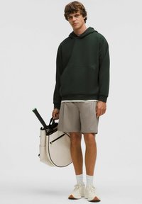 Mørkegrøn hættetrøje, grå shorts, hvide sneakers, holder en beige sportstaske med sorte detaljer og en tennisketsjer.