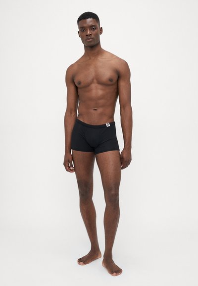 Schwarze Boxershorts aus glattem Stoff, mit einem weichen Bund und einem kleinen weißen Logo. Das Model hat einen atletischen Körperbau und steht barfuß.