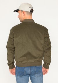 Veste bomber en velours côtelé vert olive avec des poignets et un col côtelés, dotée d'une poche latérale, portée avec un jean bleu. Vue arrière affichée.