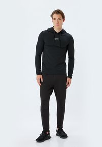 Sudadera negra de manga larga con capucha, con un bolsillo frontal y un diseño de texto ligero; combinada con pantalones ajustados negros y zapatillas deportivas negras.