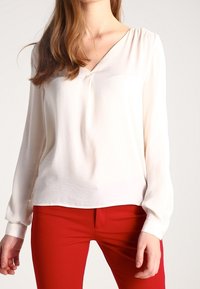Una donna indossa una blusa bianca a maniche lunghe con scollo a V abbinata a pantaloni rossi aderenti su uno sfondo neutro.