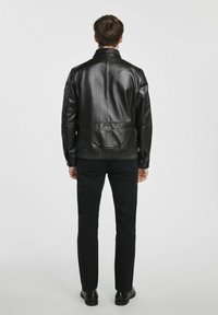 Czarna skórzana kurtka z wysokim kołnierzem, prążkowanym pasem w talii i dopasowanym krojem, noszona na ciemnych spodniach i czarnych butach. Widok z tyłu.