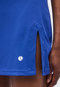 Κοντινή λήψη ενός μπλε υφασμάτινου φόρεματος με πλευρικό σκίσιμο και μικρό λευκό λογότυπο κοντά στο τελείωμα, που φοριέται από άτομο με σκουρόχρωμο δέρμα.