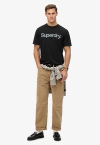 Sort bomulds T-shirt med "Superdry." logo, beige kord-agtige bukser, grå sweater bundet om taljen, og sorte loafers.