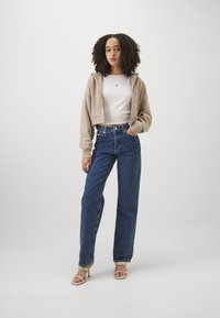 Haut à capuche beige raccourci, chemise blanche ajustée, jean bleu taille haute et sandales de couleur claire. Tissu doux avec une coupe droite.