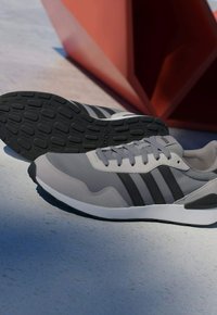 Baskets Adidas grises et noires posées sur un béton clair, une chaussure montrant la semelle texturée, avec une structure géométrique rouge en arrière-plan.
