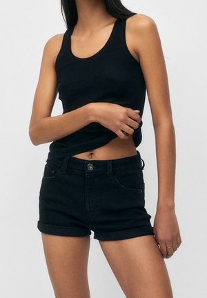 Kvinde iført sort ærmeløs top og sorte denimshorts med oprullede kanter, med den ene hånd løftende sin top let.