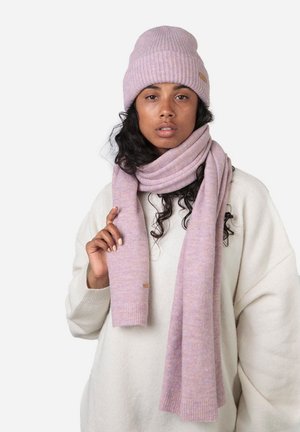 Bonnet en tricot à côtes lilas clair et écharpe assortie, fabriqués en matériau doux, portés par-dessus un pull écru, arborant une discrète étiquette de marque.