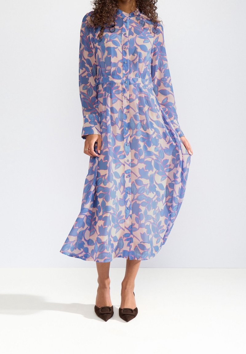 Femme portant une robe midi à manches longues boutonnée avec un motif de feuilles bleues et roses, associée à des escarpins à bout pointu de couleur foncée.